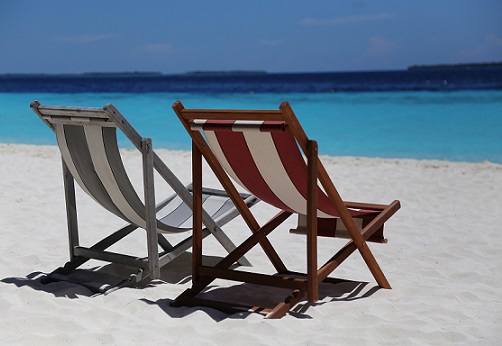 One red and white striped deck chair sits next to a grey and white striped deck chair on a white sand beach in front of a turquoise ocean fading to navy in the distance