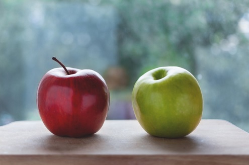 A red apple sits next to a green apple on a wooden block in front of an out of focus window