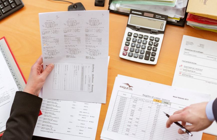 Invoices, statements and a calculator sit on a busy desk as two colleagues confer