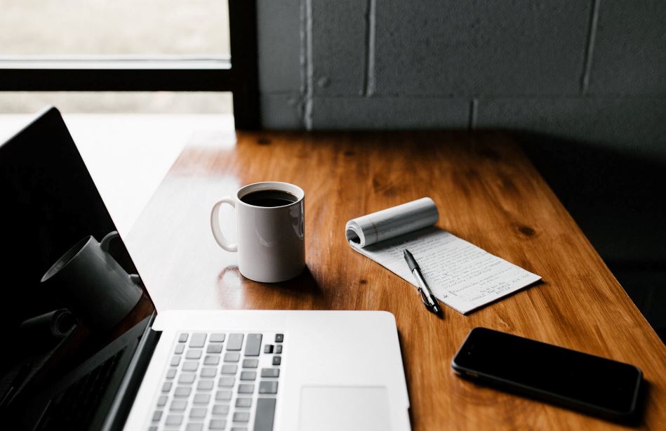 Dark colour wood desk butted up to breeze block wall and modern window, an open laptop, mobile, notepad and full coffee cop take centre stage