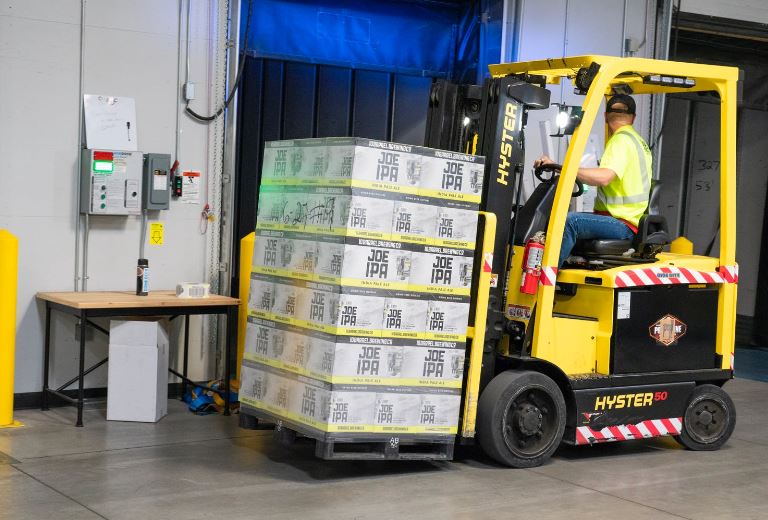 Reversing yellow forklift carries shrink wrapped pallet of boxes passed electrical control panel