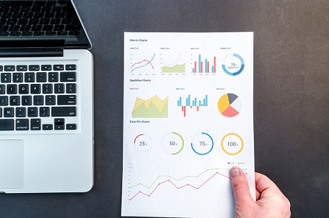 A page showing multiple types of graph and charts is placed on a dark colour desk next to a silver opened laptop