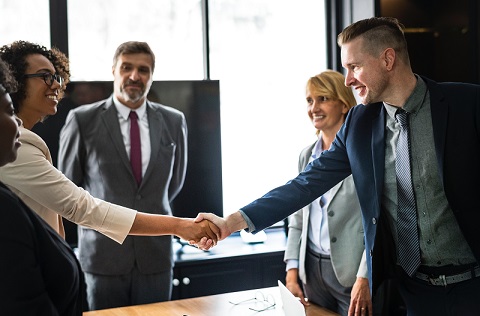 Two employees shake hands over a desk surrounded by colleagues