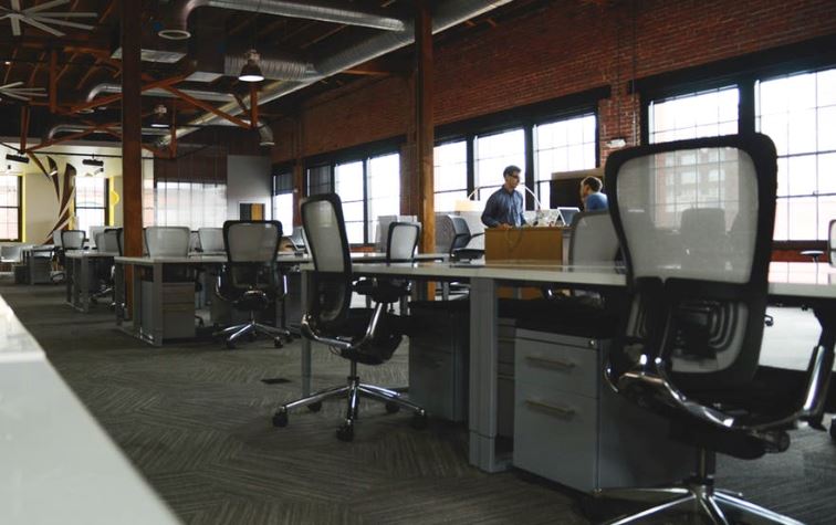 Open plan office in converted industrial space with many empty chairs and desks but two colleagues conferring