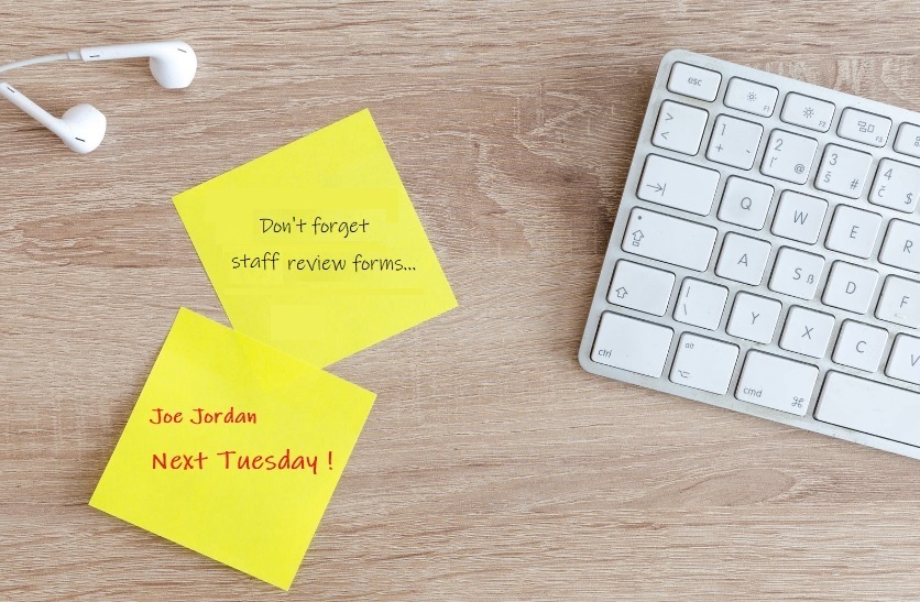 Light coloured wooden desk with earphones and apple keyboard, two yellow post notes with reminders