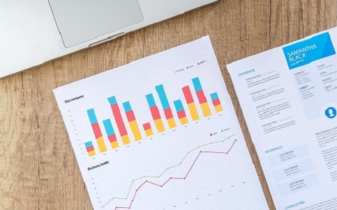 A report with two charts and a document sit on a wooden desk in front of a laptop