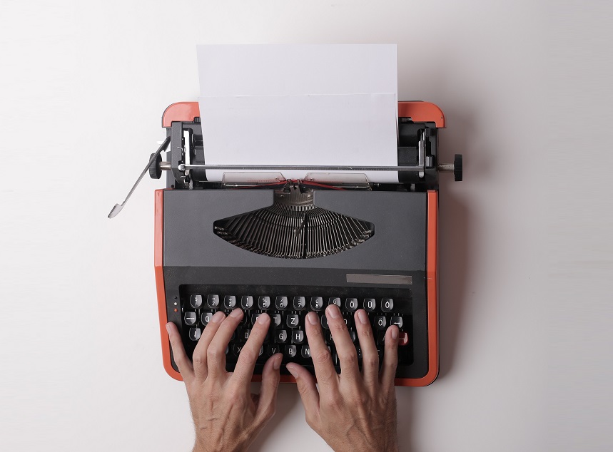 Two hands on the keyboard of a black and red type writer with a blank sheet of paper loaded