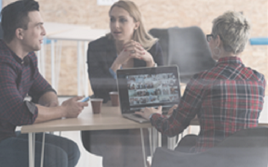 Three employees discuss around a table with coffee and an open laptop